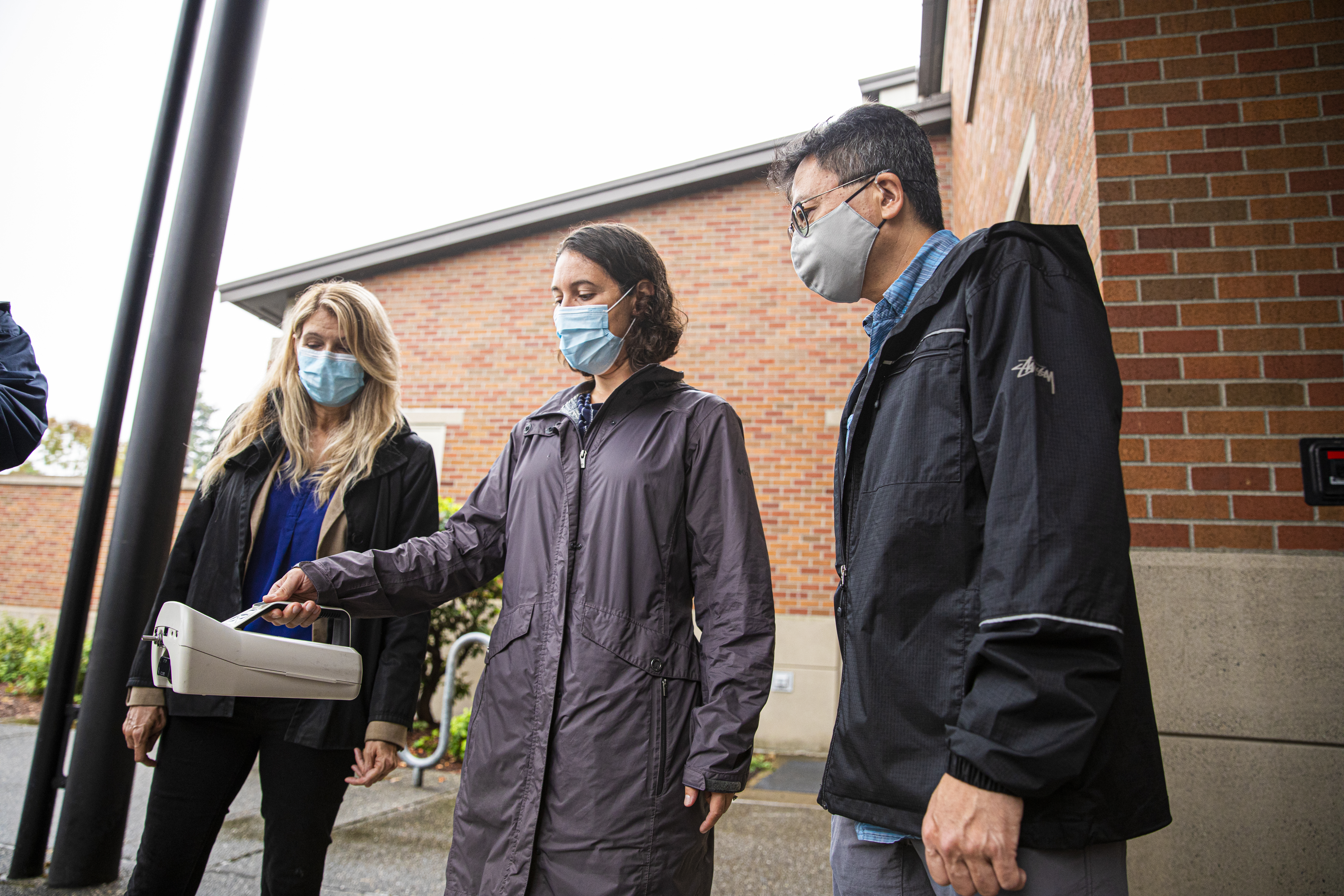 Three researchers standing side-by-side. The researcher in the middle is holding a air sampling device